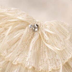 May include: A cream-colored, ruffled lace collar featuring a delicate ribbed texture. The accessory is adorned with a thin satin ribbon tied in a bow at the center, accented by a single white faux pearl and a clear, faceted crystal embellishment.