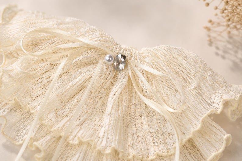 May include: A cream-colored, ruffled lace collar featuring a delicate ribbed texture. The accessory is adorned with a thin satin ribbon tied in a bow at the center, accented by a single white faux pearl and a clear, faceted crystal embellishment.