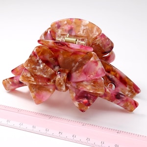 Pu&ograve; includere: Un fermaglio per capelli con motivo marmorizzato nei toni del rosa, arancione e bianco e un design a forma di fiocco. Il fermaglio &egrave; posizionato accanto a un righello rosa che mostra una larghezza di circa 10 cm nel punto pi&ugrave; ampio.