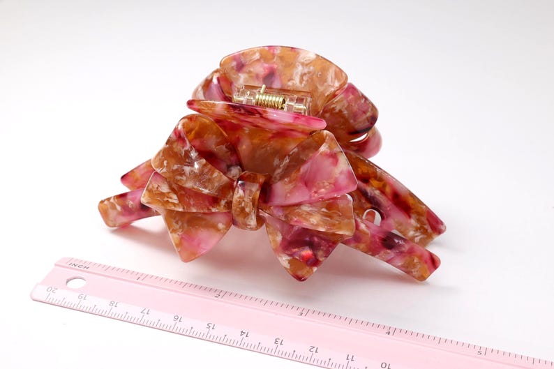 Pu&ograve; includere: Un fermaglio per capelli con motivo marmorizzato nei toni del rosa, arancione e bianco e un design a forma di fiocco. Il fermaglio &egrave; posizionato accanto a un righello rosa che mostra una larghezza di circa 10 cm nel punto pi&ugrave; ampio.