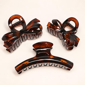 May include: Three hair claw clips with a brown and black tortoiseshell pattern. Two clips feature a bow-shaped design with interlocking teeth, while the third clip is a long, curved rectangular shape with a row of teeth along the bottom edge.
