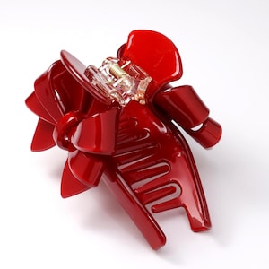 Pu&ograve; includere: Un fermaglio per capelli rosso lucido con un design a fiocco decorativo sulla parte superiore. L'accessorio &egrave; dotato di un meccanismo a cerniera in plastica trasparente e di una molla metallica centrale per fissare le ciocche di capelli.