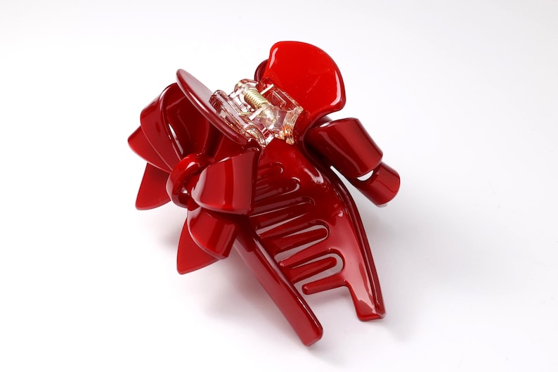Pu&ograve; includere: Un fermaglio per capelli rosso lucido con un design a fiocco decorativo sulla parte superiore. L'accessorio &egrave; dotato di un meccanismo a cerniera in plastica trasparente e di una molla metallica centrale per fissare le ciocche di capelli.