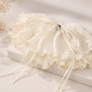 May include: A cream-colored lace garter featuring a multi-layered ruffled design with delicate scalloped edges. The accessory is adorned with a thin satin ribbon tied in a bow at the center, accented by a small white faux pearl and a clear crystal.