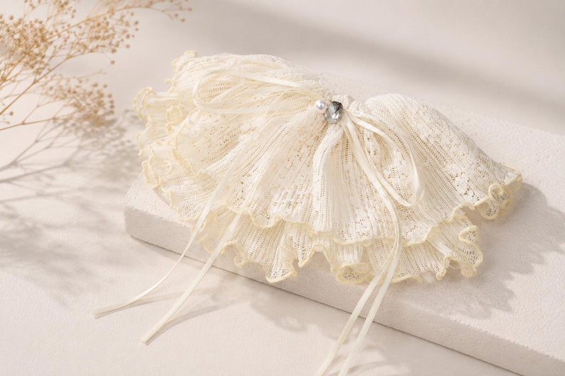 May include: A cream-colored lace garter featuring a multi-layered ruffled design with delicate scalloped edges. The accessory is adorned with a thin satin ribbon tied in a bow at the center, accented by a small white faux pearl and a clear crystal.
