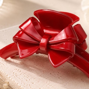 Pu&ograve; includere: Un fermaglio per capelli a artiglio di colore rosso brillante e lucido, caratterizzato da un prominente design a fiocco scultoreo sulla parte superiore. L'accessorio presenta una finitura liscia e riflettente e un meccanismo a molla funzionale.