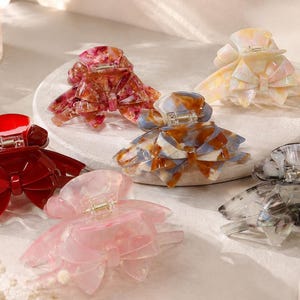 Pu&ograve; includere: Una collezione di sei mollette per capelli a forma di fiocco. Le clip sono disponibili in varie finiture, tra cui rosso tinta unita, rosa traslucido, marmorizzato blu e marrone, rosso marmorizzato, grigio marmorizzato e giallo pallido iridescente.