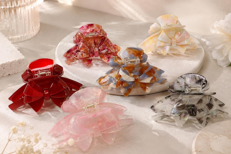 Pu&ograve; includere: Una collezione di sei mollette per capelli a forma di fiocco. Le clip sono disponibili in varie finiture, tra cui rosso tinta unita, rosa traslucido, marmorizzato blu e marrone, rosso marmorizzato, grigio marmorizzato e giallo pallido iridescente.