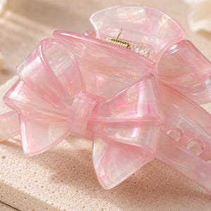 Pu&ograve; includere: Un fermaglio per capelli traslucido rosa chiaro con un design a fiocco prominente sulla parte superiore. Il materiale ha una finitura perlata e marmorizzata con sottili variazioni di colore, e un meccanismo a molla metallico &egrave; visibile sul cardine.