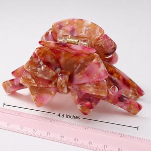Pu&ograve; includere: Un fermaglio per capelli con motivo marmorizzato nei toni del rosa, arancione e bianco, caratterizzato da un design a fiocco e un meccanismo a molla in metallo. Un righello rosa &egrave; posizionato davanti al fermaglio, indicando una larghezza di 10,9 cm.