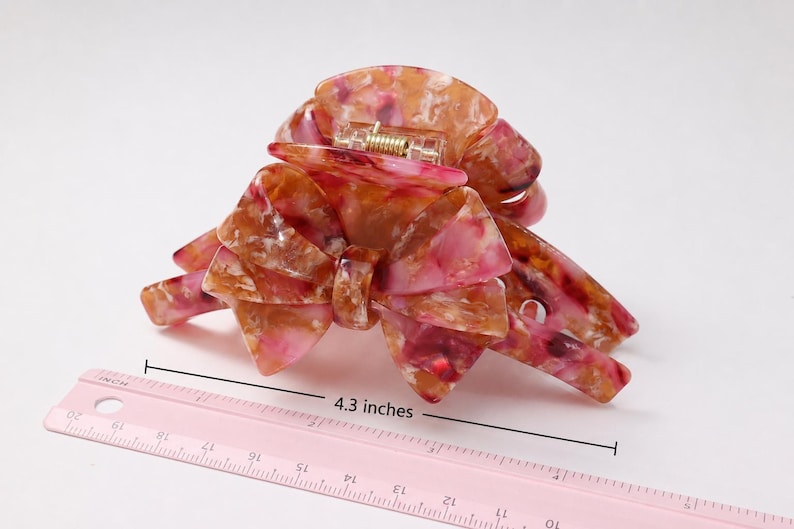Pu&ograve; includere: Un fermaglio per capelli con motivo marmorizzato nei toni del rosa, arancione e bianco, caratterizzato da un design a fiocco e un meccanismo a molla in metallo. Un righello rosa &egrave; posizionato davanti al fermaglio, indicando una larghezza di 10,9 cm.