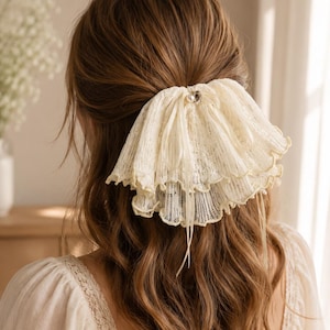 May include: A cream-colored, multi-layered lace hair accessory featuring a ruffled design with delicate scalloped edges. The accessory is centered with a small, round white pearl accent and thin ribbons hanging down from the gathered fabric base.