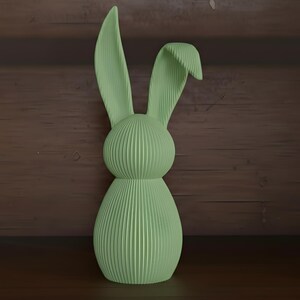 May include: A light green decorative bunny statue with a ribbed texture. The bunny has long ears and a rounded body. The statue is made of a solid material and is displayed against a wooden background.