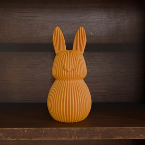 May include: An orange, textured rabbit figurine with vertical lines, standing on a wooden shelf. The bunny has long ears and a rounded body, and is set against a dark wood background.