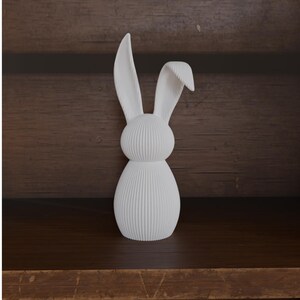 May include: A white decorative bunny figurine with a textured, ribbed design. The bunny has long ears and a rounded body. The figurine is displayed on a dark wooden shelf against a wooden background.