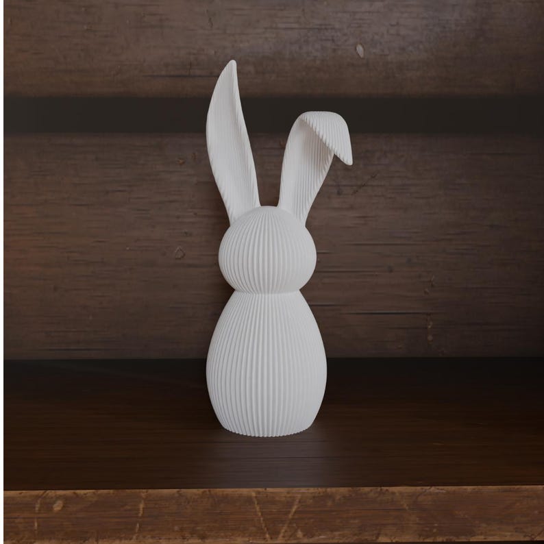 May include: A white decorative bunny figurine with a textured, ribbed design. The bunny has long ears and a rounded body. The figurine is displayed on a dark wooden shelf against a wooden background.