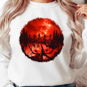 May include: White sweatshirt featuring a circular graphic with a red and black scene. The graphic depicts silhouettes of people on scooters, trees, and a stormy sky. The design has a detailed, inverted reflection.