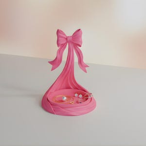 Pu&ograve; includere: Un organizer per gioielli rosa a forma di fiocco, con un vassoio circolare che contiene una collana di perle, orecchini e anelli. Il design a fiocco ha un supporto verticale e un fiocco decorativo in alto.