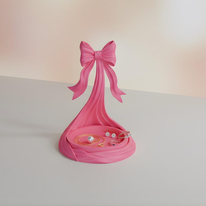 Pu&ograve; includere: Un organizer per gioielli rosa a forma di fiocco, con un vassoio circolare che contiene una collana di perle, orecchini e anelli. Il design a fiocco ha un supporto verticale e un fiocco decorativo in alto.