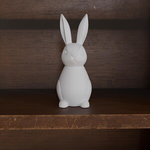 May include: A white ceramic rabbit figurine with long ears and a detailed face. The bunny is standing upright on a wooden shelf, showcasing a minimalist design. The figurine is a decorative item, perfect for Easter or spring decor.