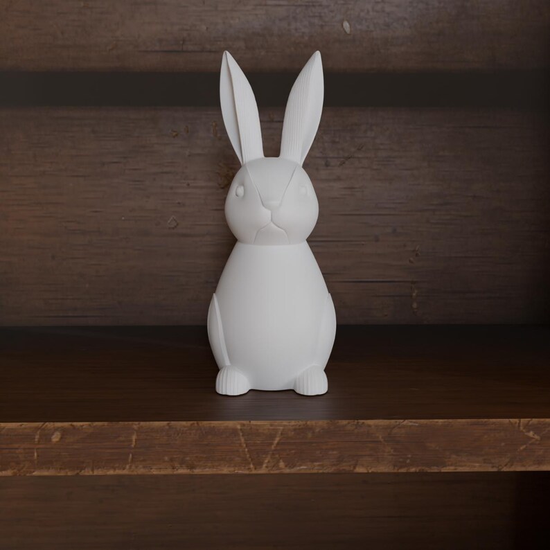 May include: A white ceramic rabbit figurine with long ears and a detailed face. The bunny is standing upright on a wooden shelf, showcasing a minimalist design. The figurine is a decorative item, perfect for Easter or spring decor.