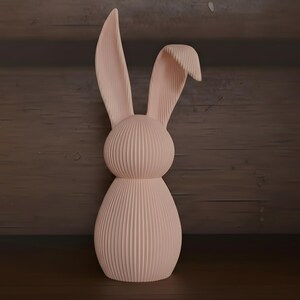 May include: A light pink bunny figurine with a ribbed texture. The bunny has long, upright ears and a rounded body. The figurine is made of a solid material and is a decorative item.