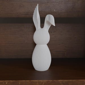 May include: A white, textured rabbit figurine with vertical lines. The rabbit has long, upright ears and a rounded body. The figurine is displayed on a dark wooden shelf, creating a contrast with the bright white color of the rabbit.