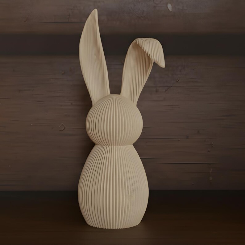 May include: A cream-colored, decorative bunny figurine. The bunny has long, upright ears and a rounded body. The surface is textured with vertical lines, giving it a ribbed appearance. The figurine is made of a solid material and stands on a wooden surface.