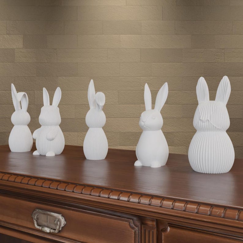 May include: Five white ceramic bunny figurines with various designs are displayed on a dark wooden surface. The bunnies have different poses and ear styles, adding a decorative touch. The background is a textured, light brown wall.