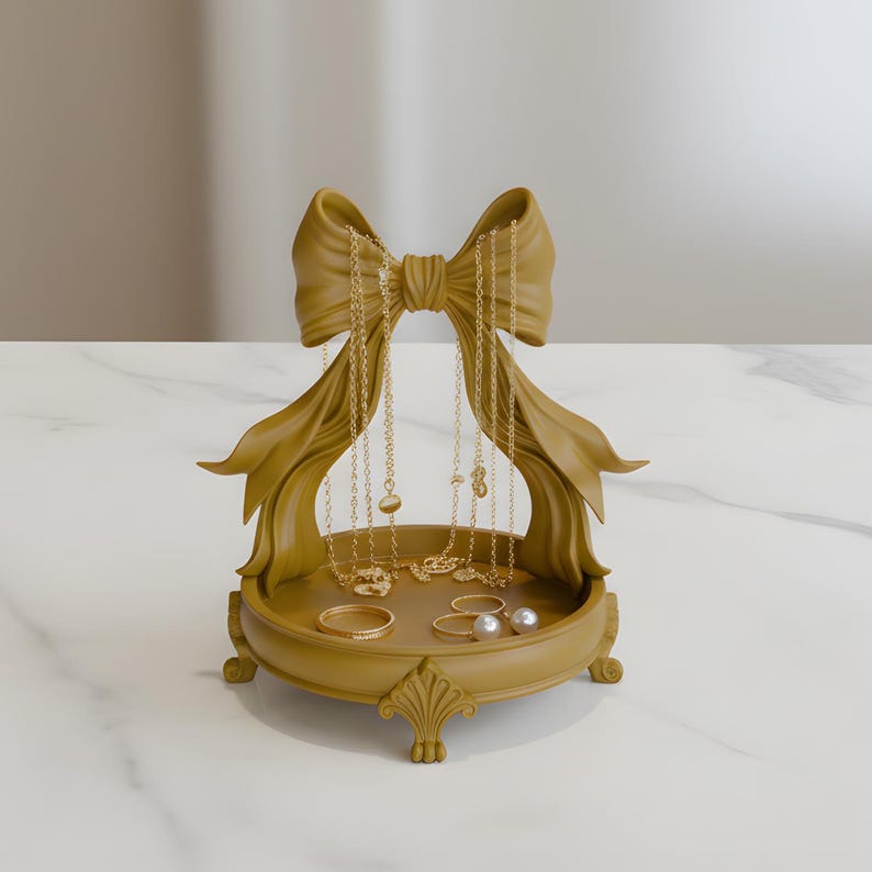 Pu&ograve; includere: Un organizer per gioielli color oro a forma di fiocco. L'organizer ha una base rotonda con un design decorativo e contiene collane, anelli e orecchini. I gioielli sono dorati e lo sfondo &egrave; una superficie simile al marmo.
