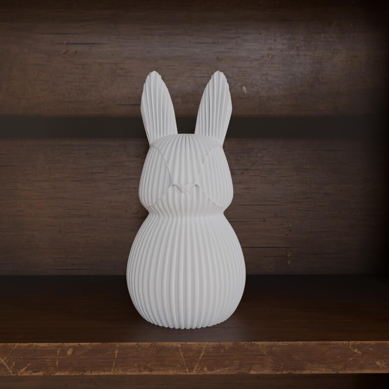 May include: A white ceramic bunny figurine with vertical ribbed detailing. The bunny has long ears and a rounded body. The figurine is displayed on a wooden shelf against a dark wood background.