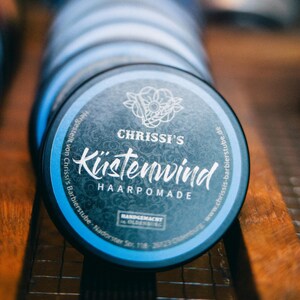 Może przedstawiać: Zbliżenie na stos okrągłych puszek z pomadą do włosów. Górna puszka jest niebiesko-czarna z napisem "CHRISSI'S Küstenwind HAARPOMADE". Puszki znajdują się na drewnianej powierzchni. Produkt jest ręcznie robiony.