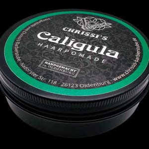 Puede incluir: Un recipiente redondo negro de pomada capilar Caligula de Chrissi's. La etiqueta es negra con texto blanco y un borde verde. El texto incluye "HANDGEMACHT in OLDENBURG" y "www.chrissis-barbierstube.de".