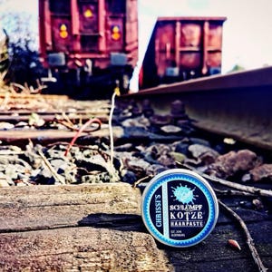 Puede incluir: Una lata redonda azul y plateada de pasta para el cabello, "CHRISSIS SCHLUMPF KOTZE HAARPASTE", descansa sobre una superficie de madera desgastada junto a una vía de tren. El fondo presenta vagones de tren rojo óxido y un lecho de grava.