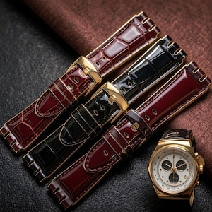 May include: Three watch straps in burgundy and black with gold-coloured buckles. The straps have a textured, crocodile-like pattern. A gold-coloured watch with a white face and black sub-dials is also present.