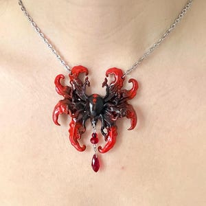 以下が含まれることがあります： 赤と黒のアクセントが施された、黒いクモのペンダントが特徴的なユニークなネックレス。赤いティアドロップ型の宝石が揺れています。シルバーチェーンに繋がれています。個性的なジュエリーです。