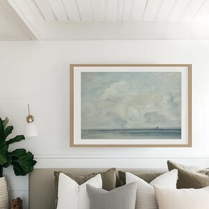 May include: A bedroom with a white bed, a white headboard, and a framed painting of a seascape above the bed. The painting is in a light wood frame. The bed has a white duvet and several pillows in shades of gray, beige, and green.