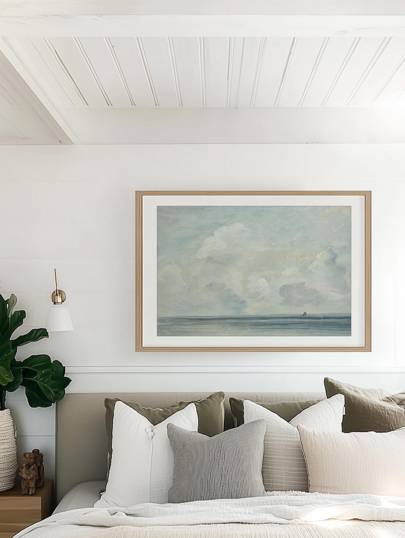 May include: A bedroom with a white bed, a white headboard, and a framed painting of a seascape above the bed. The painting is in a light wood frame. The bed has a white duvet and several pillows in shades of gray, beige, and green.