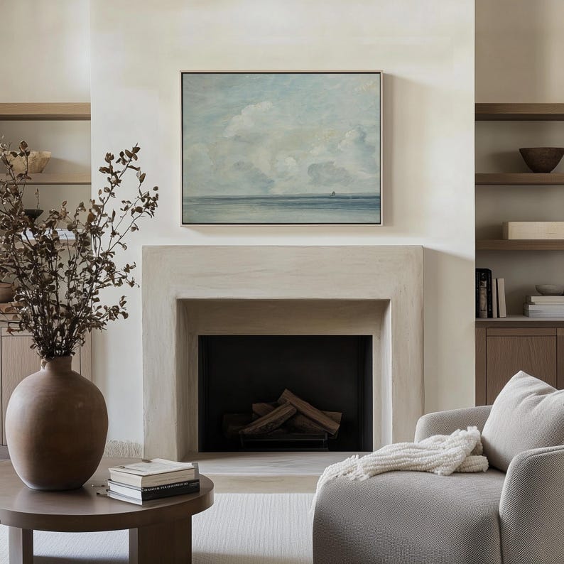 May include: A white fireplace with a black firebox and a painting of a seascape above it. The painting is framed in a light wood frame. There is a vase with dried branches on the left side of the fireplace and a white armchair on the right side.