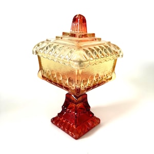 May include: A square glass candy dish with a lid, featuring a gradient colour transition from deep red at the base to a lighter amber yellow at the top. The piece has a textured, pressed glass pattern with a tiered lid and a pedestal base.