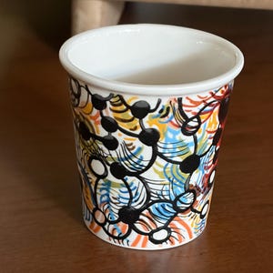 May include: A white ceramic cup with a colorful abstract design. The design features black, orange, blue, yellow, and white shapes. The cup has a smooth, glossy finish and a simple, cylindrical shape.