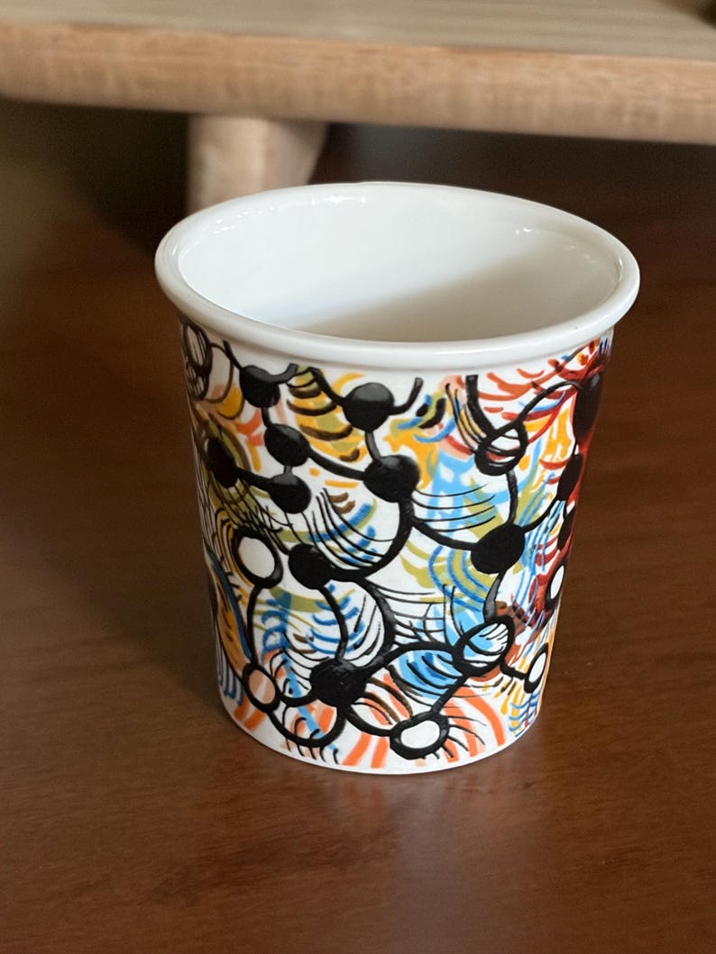 May include: A white ceramic cup with a colorful abstract design. The design features black, orange, blue, yellow, and white shapes. The cup has a smooth, glossy finish and a simple, cylindrical shape.