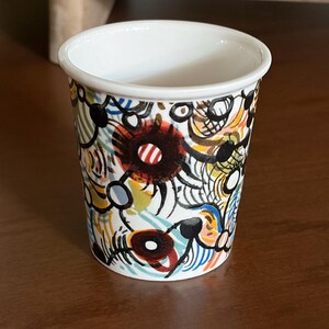 May include: A small, white ceramic cup with a colorful abstract design. The cup features a white rim and is adorned with a pattern of black, red, blue, yellow, and white shapes. The cup is sitting on a brown surface.