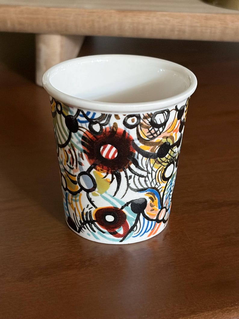 May include: A small, white ceramic cup with a colorful abstract design. The cup features a white rim and is adorned with a pattern of black, red, blue, yellow, and white shapes. The cup is sitting on a brown surface.