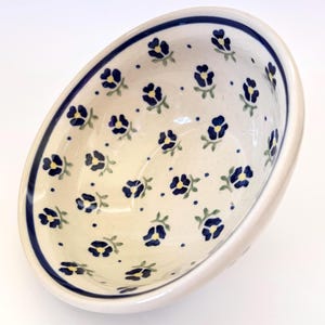 May include: A ceramic bowl with a cream-colored base featuring a repeating pattern of small dark blue flowers with yellow centers and green stems. The bowl has a thick dark blue band around the interior rim and a smooth, glossy finish throughout.