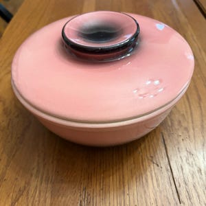 Puede incluir: Un cuenco de cerámica rosa con tapa a juego. La tapa tiene un asa negra y rosa. El cuenco y la tapa son redondos y están sobre una superficie de madera. El cuenco es probablemente para servir comida.