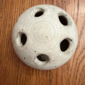 May include: A white ceramic sphere with six large, evenly spaced holes. The surface has subtle concentric circles and small dark spots. The object is likely a decorative item or a unique design element.