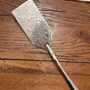 May include: A silver-toned metal cake server featuring an elongated rectangular blade with a serrated edge on one side. The surface of the blade is decorated with an intricate, symmetrical engraved floral pattern, and it has a smooth, slender handle.