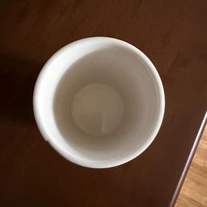 May include: A white ceramic cup with a rounded rim. The cup is empty and sits on a dark brown wooden surface. The interior of the cup is a light cream color.
