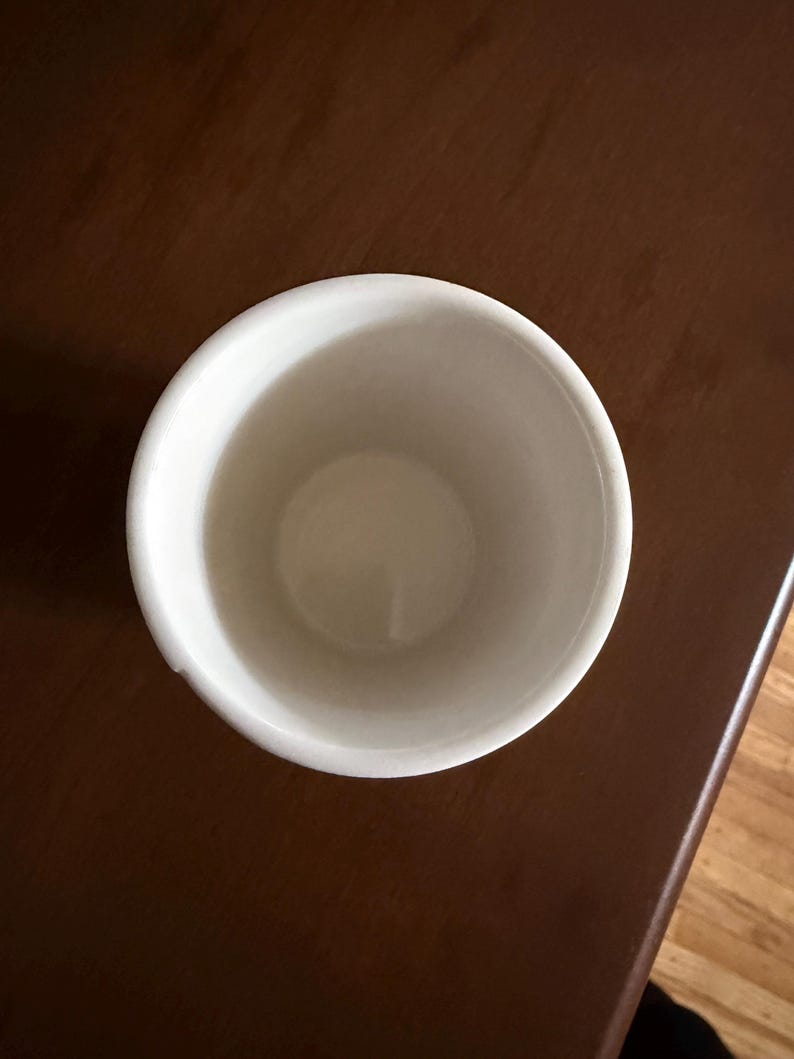 May include: A white ceramic cup with a rounded rim. The cup is empty and sits on a dark brown wooden surface. The interior of the cup is a light cream color.